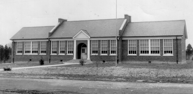 Black and white of Woodlawn Elementary School taken in 1958 for the Fairfax County School Board’s fire insurance survey of school properties. The building looks much the same as it did in the 1940s. Trees have been planted on the grounds, but they are not large yet. The building has an arched entryway between two white columns. Large windows face the street which remains unpaved. 
