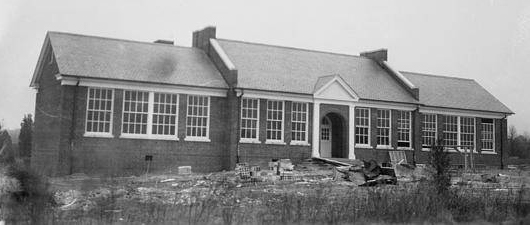 Black and white photograph of the new, brick Woodlawn Elementary School, circa 1941, during construction of two additional classrooms to the building. Construction debris is scattered about the school grounds. 
