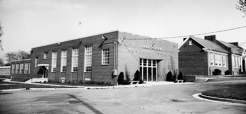 Black and white of Woodlawn Elementary School taken in 1968. Large, new sections have been added to the building. The old wing is visible on the right, a new wing stretches into the distance on the left, and in the foreground is a rectangular section with the words Auditorium, Woodlawn School, emblazoned on the building. The new and old portions of the building are very different in character. The new sections resemble other schools built in the 1950s and 60s and have a distinctly modern character compares with the portion of the school built in the 1930s.  